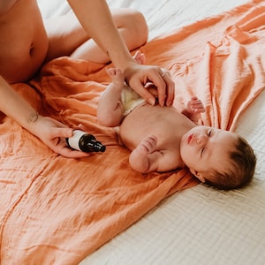 May include: A person is giving a baby a massage on a peach-colored blanket. The baby is lying on its back with its eyes closed. A small bottle of oil is on the blanket.