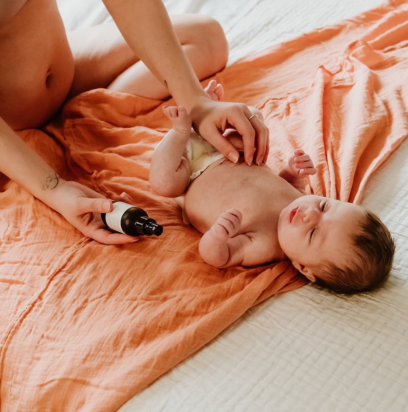 May include: A person is giving a baby a massage on a peach-colored blanket. The baby is lying on its back with its eyes closed. A small bottle of oil is on the blanket.