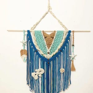 May include: A macrame wall hanging in shades of blue, turquoise, and beige, with a wooden dowel and hanging cord. The design includes fringe, decorative beads, and attached seashell and starfish accents. The wall decor is approximately 24 inches wide.