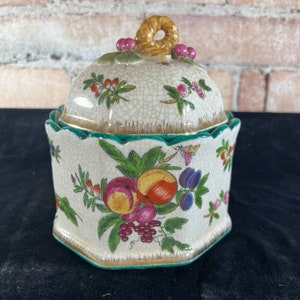 May include: A decorative ceramic jar with a lid, featuring a fruit and floral design. The jar has a cream-colored base with a cracked glaze effect and is adorned with colorful illustrations of peaches, plums, and grapes. The lid has a woven handle and pink berry accents.