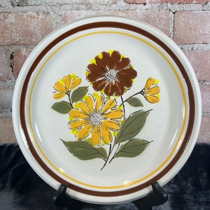 Puede incluir: Plato redondo de cerámica con diseño floral. El plato presenta un centro color crema con un estampado floral marrón y amarillo. El borde tiene un borde marrón y amarillo. El plato se exhibe sobre un soporte negro.