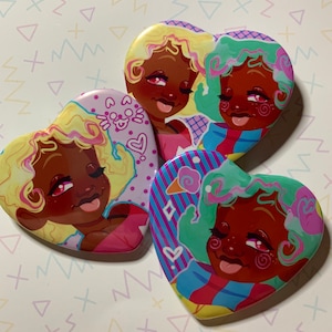 Puede incluir: Tres pines en forma de corazón con fondos coloridos e ilustraciones de personajes de dibujos animados con piel marrón y cabello colorido. Cada pin presenta un personaje diferente con una expresión y un peinado únicos.