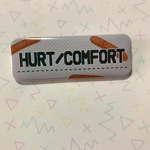 Może przedstawiać: Biały guzik z szarym tłem i kropkowanym wzorem. Guzik ma wz&oacute;r brązowego bandaża i napis "HURT/COMFORT" w kolorze zielonym.