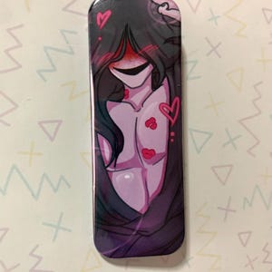 Puede incluir: Pin rectangular con una ilustración estilizada de una figura con pelo largo negro, ojos rojos y una amplia sonrisa. El pecho de la figura está adornado con corazones y labios rosas. El fondo es un degradado de morado y negro.