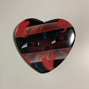 May include: Heart-shaped button with a cartoon design. The button features a black background with red and pink accents, including hearts and stylized eyes. The design has a cute, anime-inspired aesthetic.