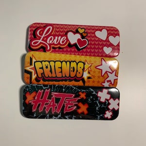 Puede incluir: Tres pinzas de pelo rectangulares de metal. La pinza superior es rosa con la palabra "Love" en escritura roja y corazones blancos. La pinza del medio es naranja con la palabra "FRIENDS" en amarillo. La pinza inferior es negra con la palabra "HATE" en rojo.