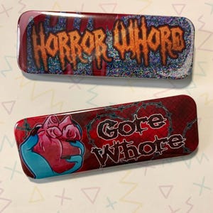 Puede incluir: Dos pines rectangulares rojos y negros con un acabado brillante. El pin superior dice "Horror Whore" en letras naranjas. El pin inferior dice "Gore Whore" en letras negras con un diseño de alambre de púas y una mano azul sosteniendo un corazón rojo.