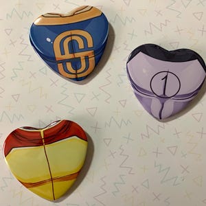 Op de afbeelding: Drie hartvormige pinnen met verschillende ontwerpen. De eerste pin is blauw met een gouden 'S' erop. De tweede pin is paars met een witte '1' erop. De derde pin is geel met rode en oranje accenten.