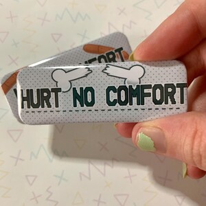 Może przedstawiać: Biały guzik z czarno-białym wzorem kości i tekstem "Hurt No Comfort" w zielonych literach.