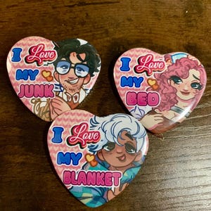 May include: Three heart-shaped buttons with illustrations and text. Each button features a different character and the phrase "I Love My" followed by "Junk," "Bed," and "Blanket." The buttons have a pink background with a heart pattern.