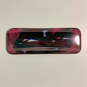 Puede incluir: Una lata de metal rectangular con un diseño de ojo estilizado. La obra de arte presenta dos ojos con acentos rojos, reflejos azules y un fondo oscuro con tonos rosas y verdes. La lata tiene esquinas redondeadas.