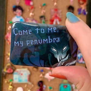 May include: A rectangular, dark blue charm with the text "Come to me, my penumbra" in white. The charm features a shadowy figure with glowing white eyes. Other charms are visible in the background.