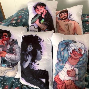 May include: A collection of white rectangular throw pillows, each featuring a colorful anime-style illustration of a person. The illustrations depict various characters with unique hairstyles, clothing, and expressions. The pillows are arranged on a patterned blanket.