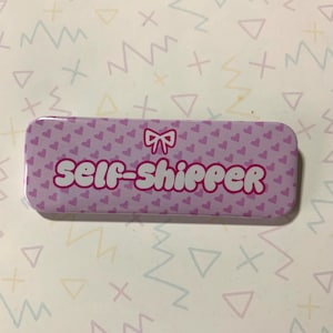 May include: A pink button with a white background and a pink bow. The button has a pink heart pattern and the text "self-shipper" in white.
