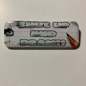 May include: Rectangular art-themed button with the text "SHUT UP AND DRAW!!" in a graffiti-style font. The design includes a pencil, an eye, and a lined paper background.