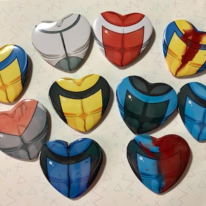 Op de afbeelding: Tien hartvormige knopen met verschillende kleurrijke ontwerpen. Elke knop toont een gestileerde menselijke torso met verschillende gekleurde kleding en patronen.