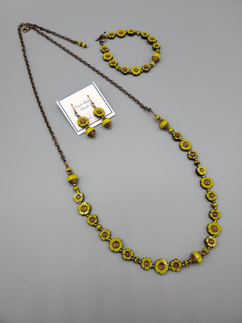 K&ouml;nnte beinhalten: Ein Set aus gelben, blumenf&ouml;rmigen tschechischen Glasperlen. Das Set enth&auml;lt eine Halskette, ein Armband und Ohrringe. Die Perlen sind auf einer Bronzekette aufgef&auml;delt.