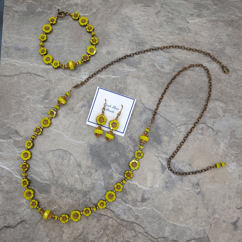 K&ouml;nnte beinhalten: Ein Set aus gelben Glasblumenperlen-Schmuck, bestehend aus einer Halskette, einem Armband und Ohrringen. Die Halskette und das Armband bestehen aus einer Kette aus kleinen, gelben Glasblumen. Die Ohrringe sind schlicht, mit jeweils einer einzelnen gelben Glasblume.