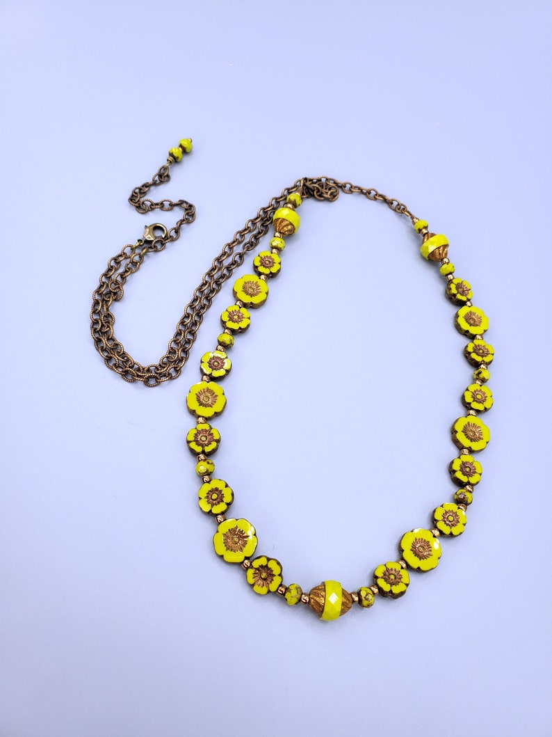 K&ouml;nnte beinhalten: Eine gelbe Blumenkette mit einer goldenen Kette. Die Kette besteht aus Glasperlen mit einem Blumenmuster.