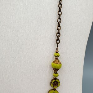 K&ouml;nnte beinhalten: Eine lange Halskette mit einer Messingkette und gr&uuml;nen Glasperlen. Die Perlen sind blumenf&ouml;rmig und haben eine goldfarbene Oberfl&auml;che.