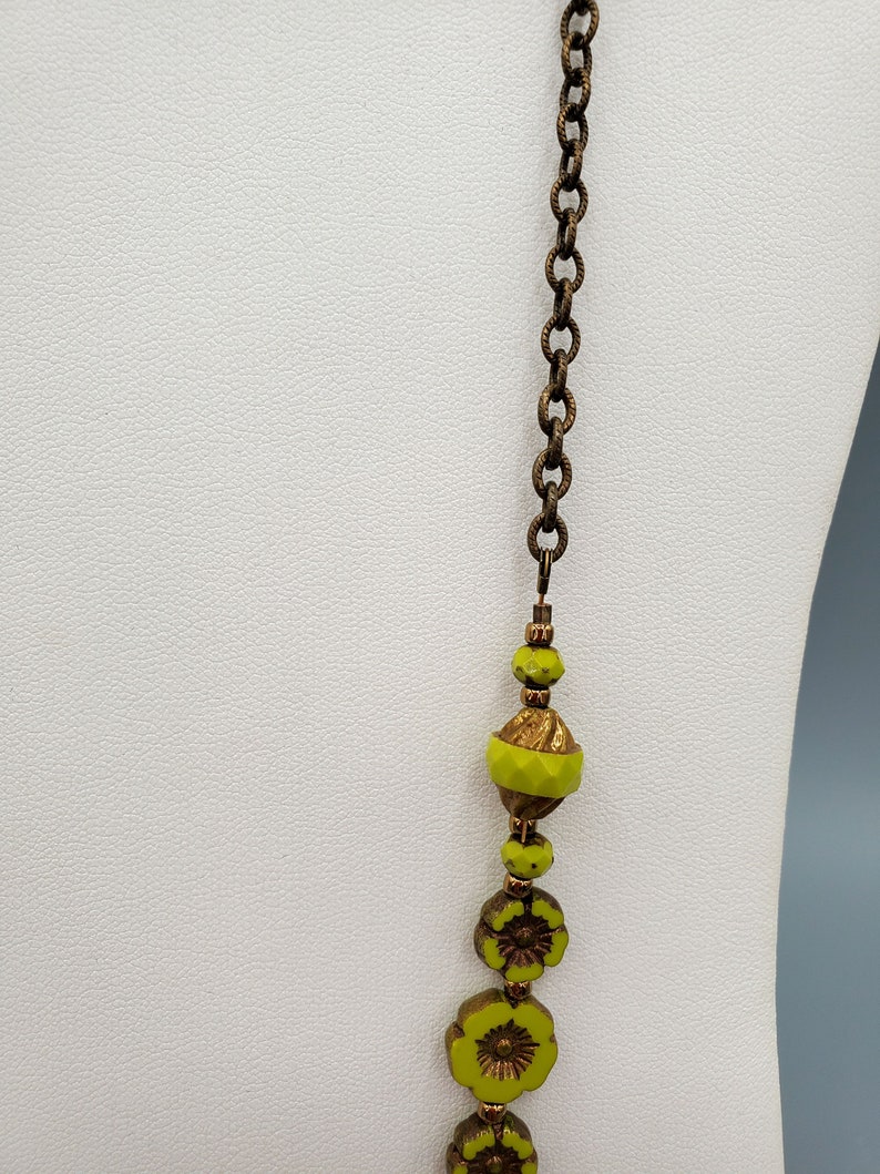 K&ouml;nnte beinhalten: Eine lange Halskette mit einer Messingkette und gr&uuml;nen Glasperlen. Die Perlen sind blumenf&ouml;rmig und haben eine goldfarbene Oberfl&auml;che.