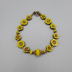 K&ouml;nnte beinhalten: Ein gelbes Blumenarmband mit einem Messingverschluss. Das Armband besteht aus kleinen, runden Perlen mit einem erhabenen Blumendesign.