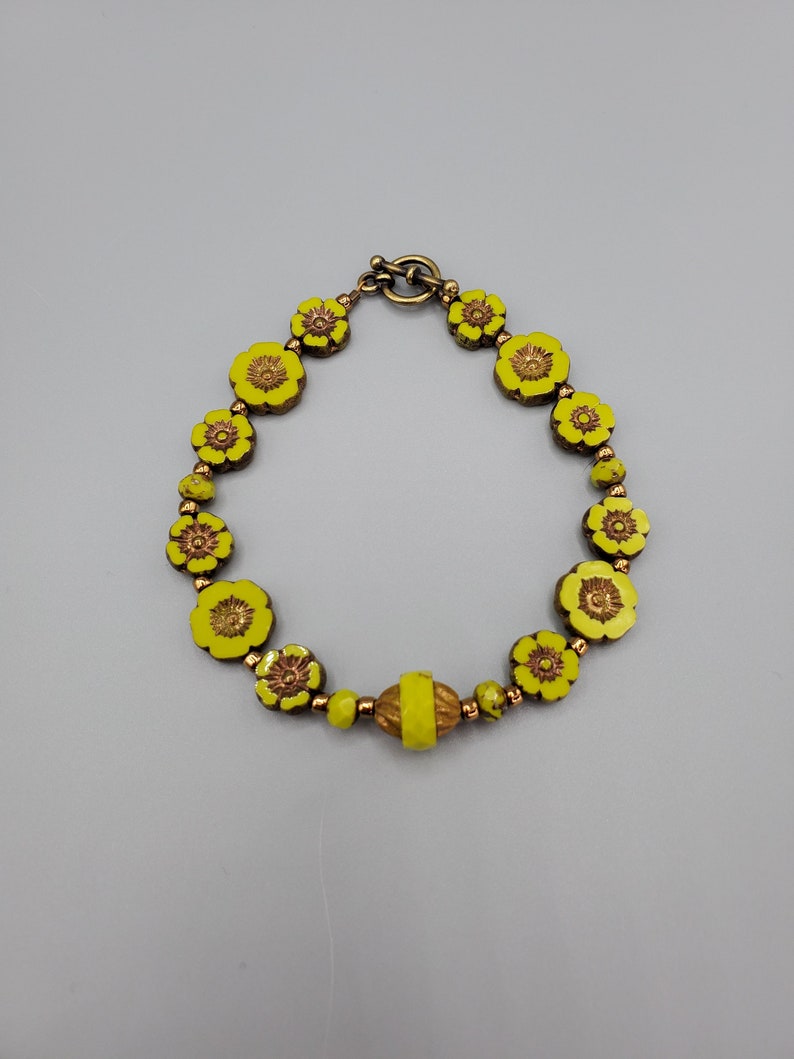 K&ouml;nnte beinhalten: Ein gelbes Blumenarmband mit einem Messingverschluss. Das Armband besteht aus kleinen, runden Perlen mit einem erhabenen Blumendesign.