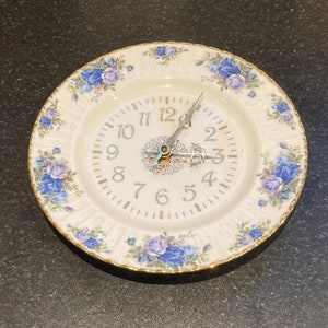 Puede incluir: Un plato de porcelana blanca con borde dorado y diseño floral azul. El plato presenta una esfera de reloj con números romanos negros y manecillas plateadas.