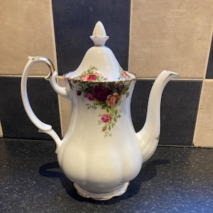 Puede incluir: Una tetera de porcelana blanca con ribete dorado y un patrón floral con rosas rojas, rosas y amarillas. La tetera tiene una tapa con un pomo en la parte superior.