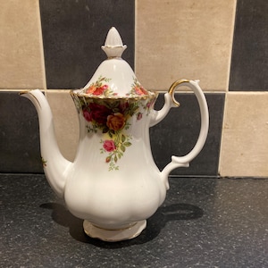 Puede incluir: Una tetera de porcelana blanca con ribete dorado y un patrón floral. La tetera tiene una tapa con un pomo en la parte superior. La tetera está sentada sobre una superficie a cuadros en blanco y negro.