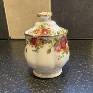 May include: White porcelain sugar bowl with a gold trim and a floral pattern featuring red, yellow, and pink roses. The bowl has a lid with a gold knob.