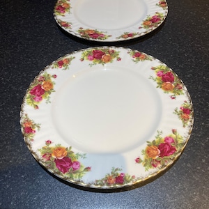 Puede incluir: Dos platos de cena de cerámica blanca con borde dorado y un patrón floral de rosas rojas, amarillas y verdes.