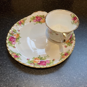 Puede incluir: Un juego de taza y platillo de té blanco con borde dorado y estampado floral. La taza tiene asa y el platillo tiene un borde festoneado.