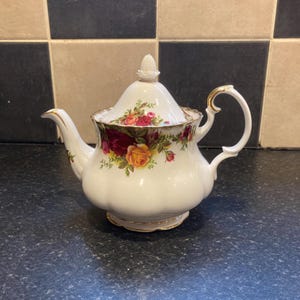 May include: White ceramic teapot with a floral rose pattern in red, yellow, and green. The teapot has a curved spout, a curved handle with gold accents, and a matching lid with a finial. The teapot is sitting on a dark surface.