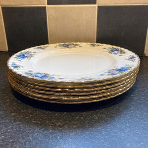Può includere: Una pila di sei piatti in ceramica bianca con un motivo floreale blu sul bordo e un bordo dorato. I piatti hanno un bordo smerlato. I piatti sono impilati su una superficie scura.
