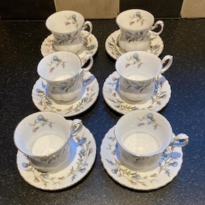 Pode incluir: Conjunto de seis chávenas e pires brancos com um design floral. Cada chávena e pires apresenta um delicado padrão floral em azul, rosa e verde, com acabamento dourado. As chávenas têm pegas. Os pires têm cerca de 14 cm de diâmetro.