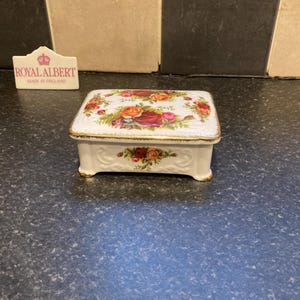 Puede incluir: Un joyero rectangular de porcelana blanca con un colorido estampado floral de rosas rojas, amarillas y rosas con hojas verdes. La tapa y la base tienen detalles en relieve y un borde dorado. Un letrero de la marca Royal Albert está cerca.