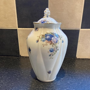 May include: A white ceramic jar with a lid, decorated with blue and pink floral patterns. The jar has a fluted design and gold trim. The lid features a decorative finial. A classic piece for home decor.