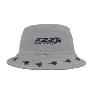 May include: A gray bucket hat with a black F22 RAPTOR logo and a black silhouette of a fighter jet repeated around the brim.