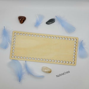 Puede incluir: Un telar de madera rectangular con agujeros alrededor de los bordes, perfecto para proyectos de tejido. El telar está sobre una superficie blanca con plumas azules y pequeñas piedras.