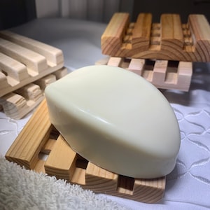 May include: A light-colored bar of soap rests on a wooden soap dish. The soap dish is made of light-colored wood with a slatted design. A white towel is in the foreground. Additional soap dishes are in the background.