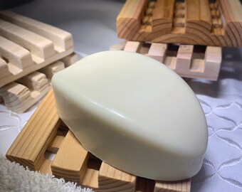 May include: A light-colored bar of soap rests on a wooden soap dish. The soap dish is made of light-colored wood with a slatted design. A white towel is in the foreground. Additional soap dishes are in the background.