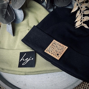 Könnte beinhalten: Zwei Strickmützen, eine olivgrün und eine schwarz, mit einem kleinen braunen Lederflicken auf der schwarzen Mütze. Der Flicken hat ein Leopardenmuster und ein kleines Herz. Die olivgrüne Mütze hat ein schwarzes quadratisches Etikett mit dem Wort "hej" in weiß mit einem kleinen Herz.