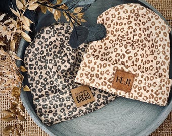 Puede incluir: Dos gorros de punto de leopardo marrón y beige con parches de cuero marrón. Un parche dice "HEJ!" y el otro dice "ciao KAKAO".
