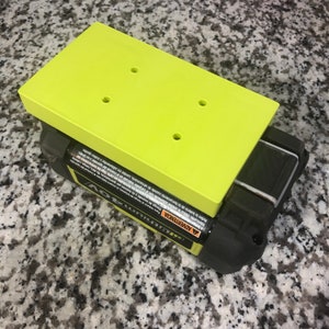 Op de afbeelding: Een groene plastic batterijhouder met vier gaten voor schroeven. De houder zit bovenop een zwart-gele batterij. De batterij heeft een waarschuwingsetiket in het Spaans en Engels.