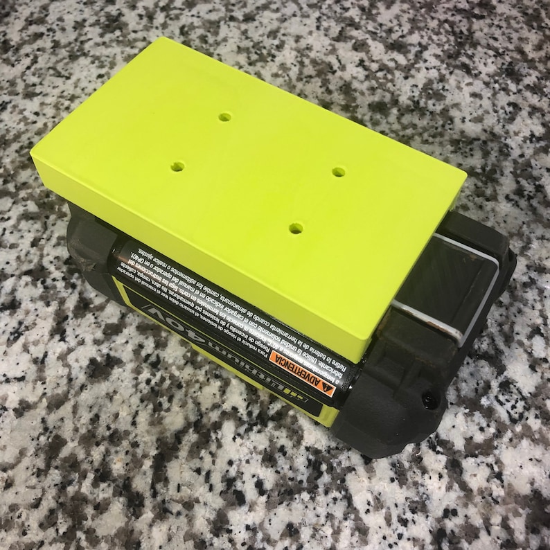 Op de afbeelding: Een groene plastic batterijhouder met vier gaten voor schroeven. De houder zit bovenop een zwart-gele batterij. De batterij heeft een waarschuwingsetiket in het Spaans en Engels.