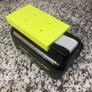 Op de afbeelding: Een geel plastic platform met vier gaten zit bovenop een zwart en grijs 40V Lithium-ion-accu. De accu heeft een witte ontgrendelingsknop.