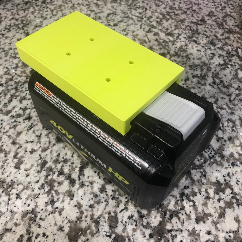 Op de afbeelding: Een geel plastic platform met vier gaten zit bovenop een zwart en grijs 40V Lithium-ion-accu. De accu heeft een witte ontgrendelingsknop.