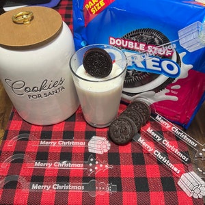 May include: A glass of milk with an Oreo cookie dipped in it, sitting on a red and black checked tablecloth. There are three clear plastic stirrers with the words "Merry Christmas" printed on them. A cookie jar with the words "Cookies for Santa" is in the background. A packet of Double Stuf Oreo cookies is also in the background.