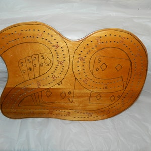 May include: A wooden cribbage board with a light brown finish. The board features a double-track design with drilled holes for pegging scores. Black ink drawings of playing cards and numbers are visible.